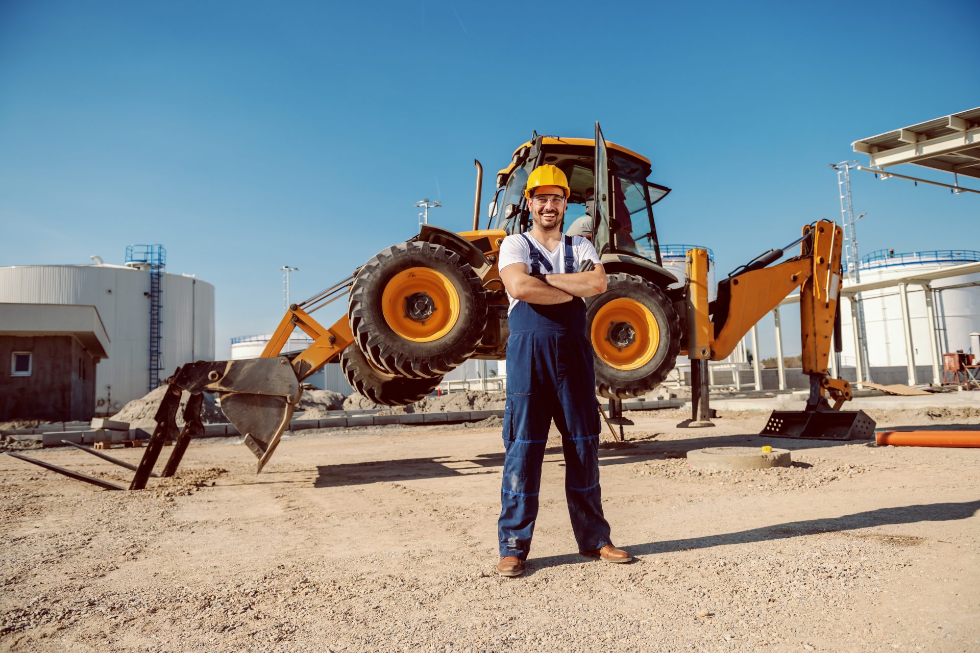 handsome-caucasian-worker-in-overall-and-helmet-on-head-standing-outdoors-with-crossed-arms